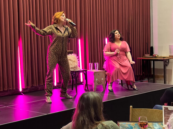Zwei Frauen, die auf einer B&uuml;hne mit rotem Vorhang im Hintergrund zu sehen sind. Eine Frau, sie hat schwarze Haare und tr&auml;gt ein pinkes Kleid, sitzt auf einem Stuhl. Daneben steht eine Frau mit orangenen Haaren und einem Leoparden-Muster-Anzug. Sie h&auml;lt ein Mikrofon vor ihr Gesicht, scheint zu singen.